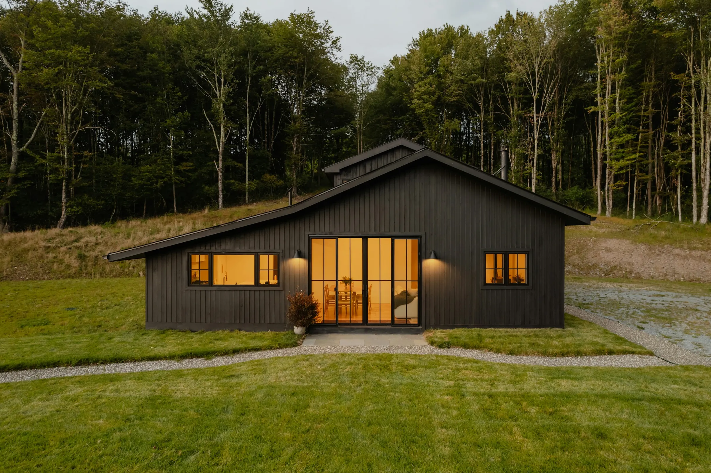 Bridleway: a long black timber house at dusk on a mown lawn, windows lit