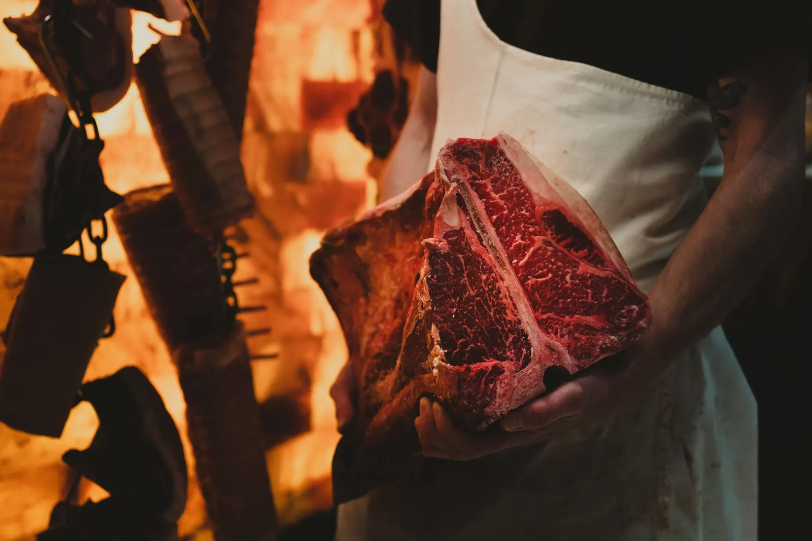 A butcher holding a marbled bone-in steak beside an open fire