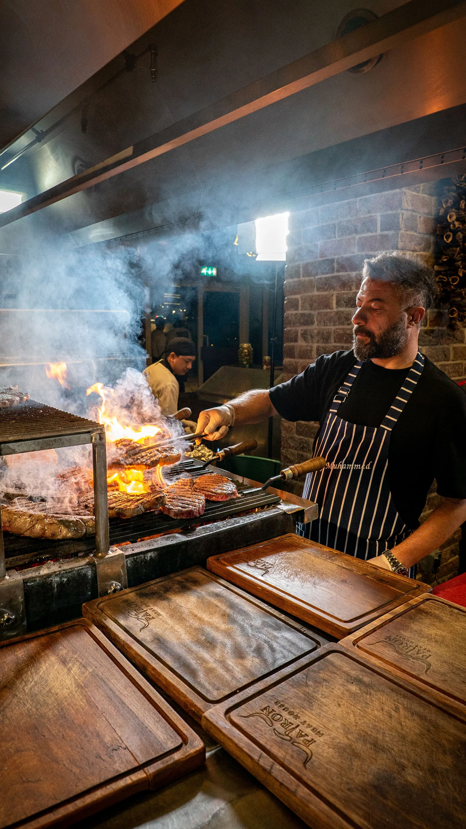 A chef in a striped apron working a smoking grill, tongs in hand