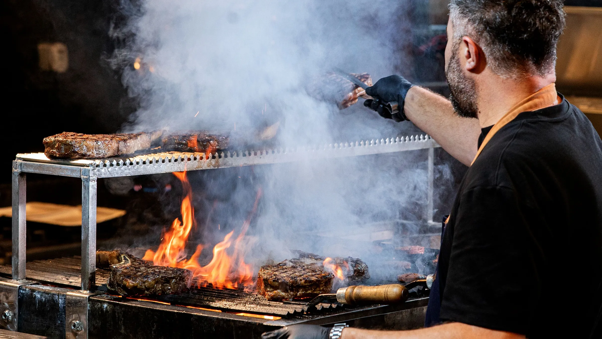 A chef pointing across a smoking grill, flames under the steaks