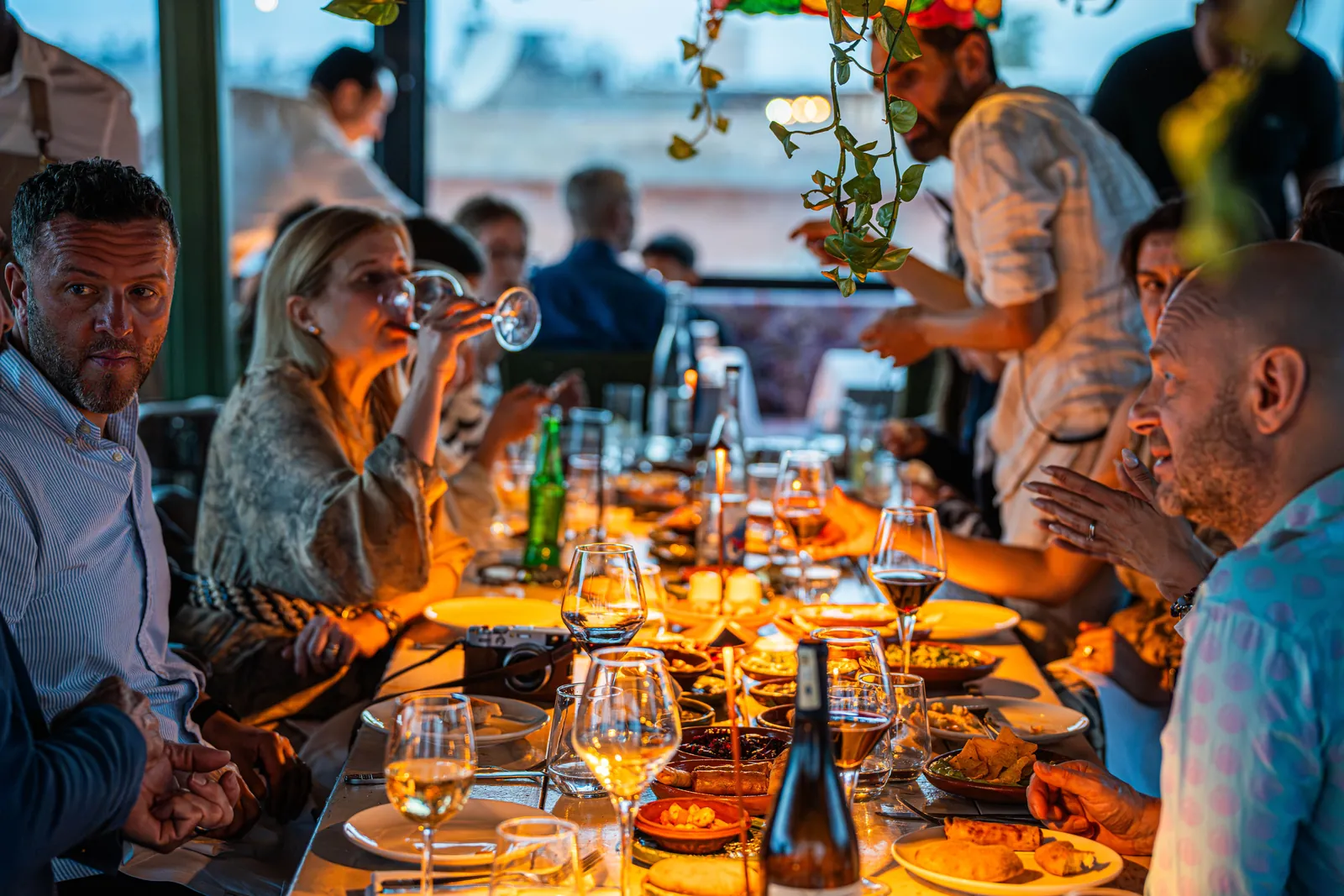 A long table of guests eating, drinking and talking under warm lights