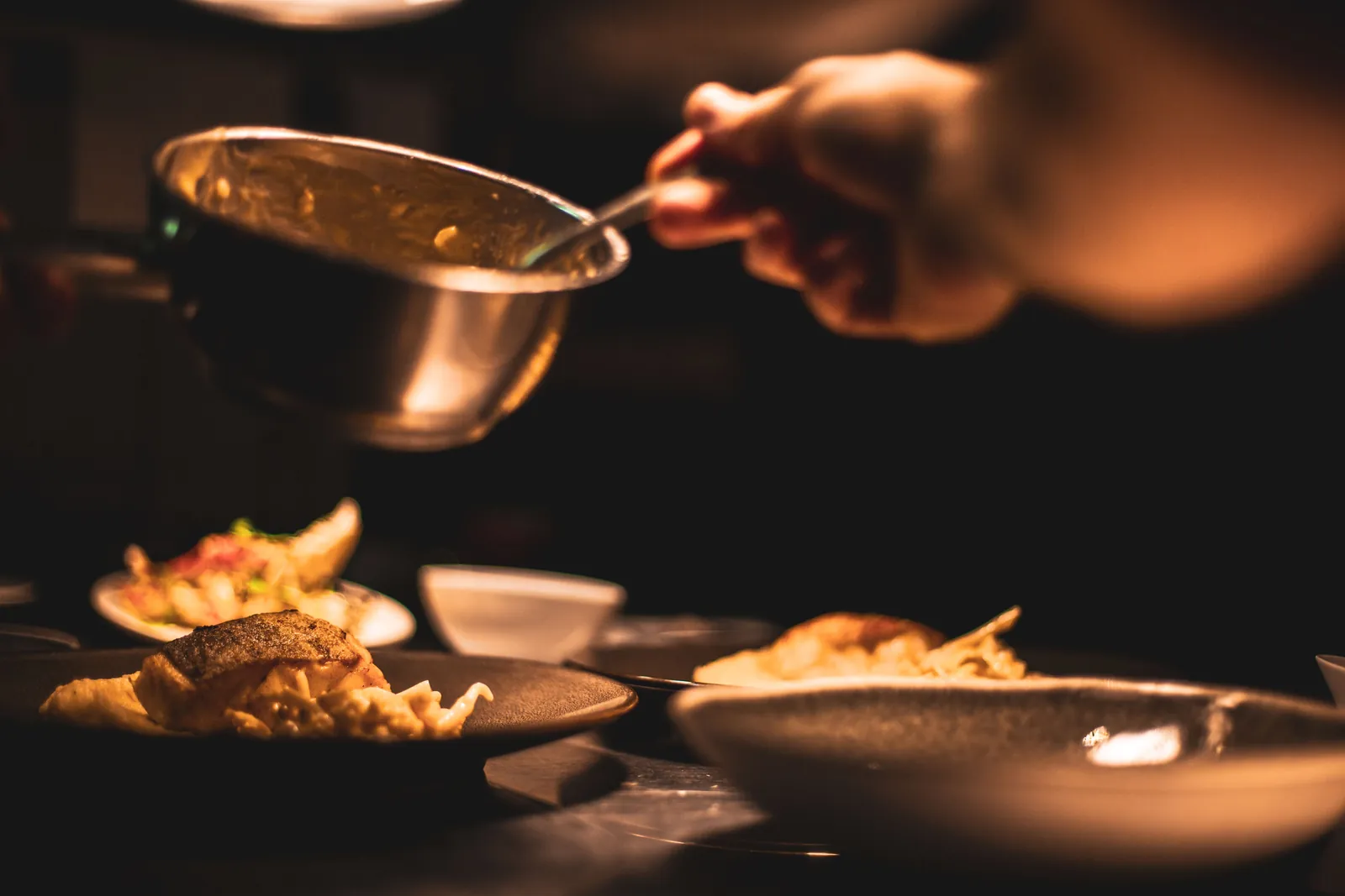 A chef spooning sauce over a plate at a dim kitchen pass