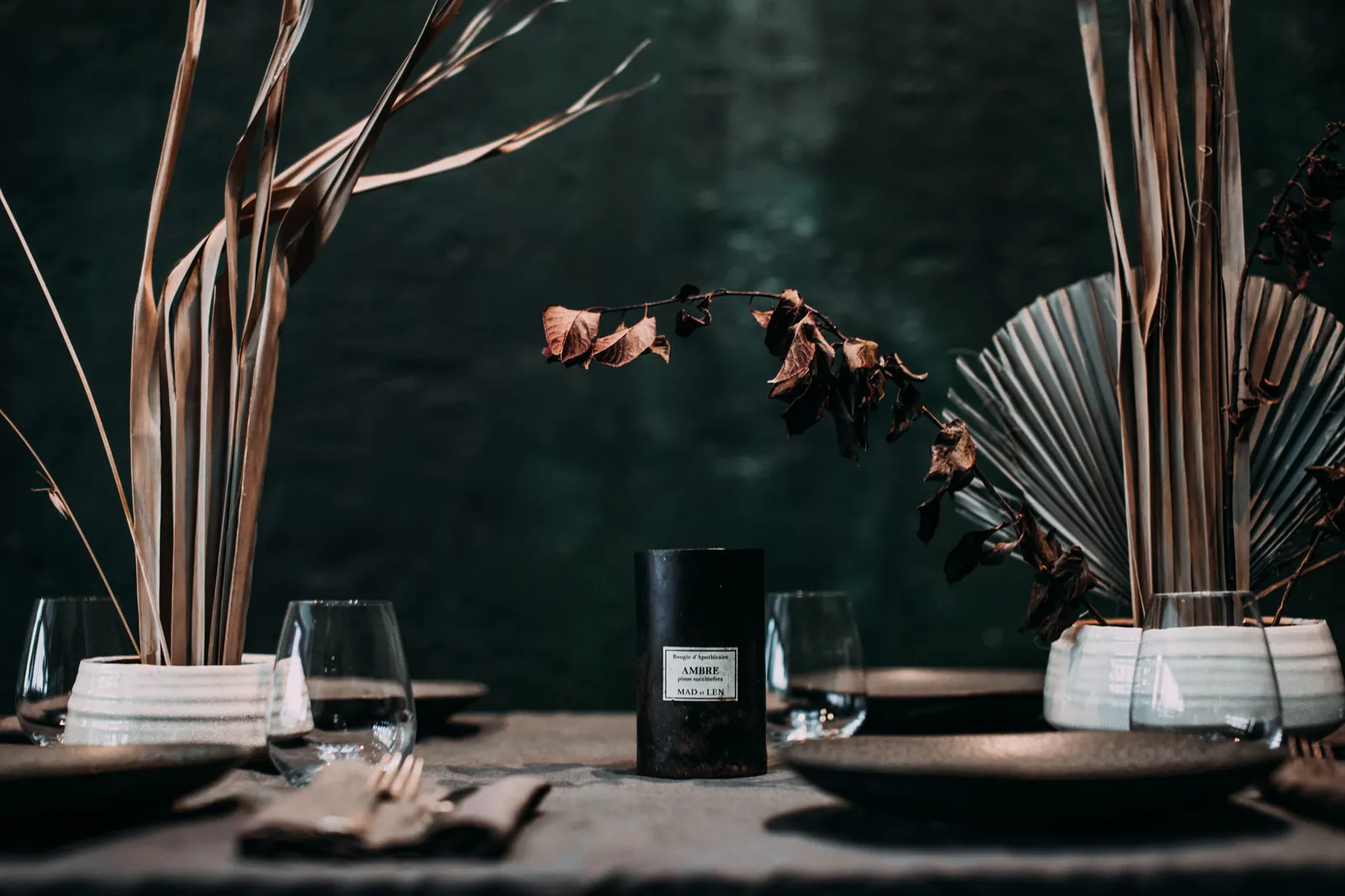 A dark table laid with black plates, glasses and dried grasses