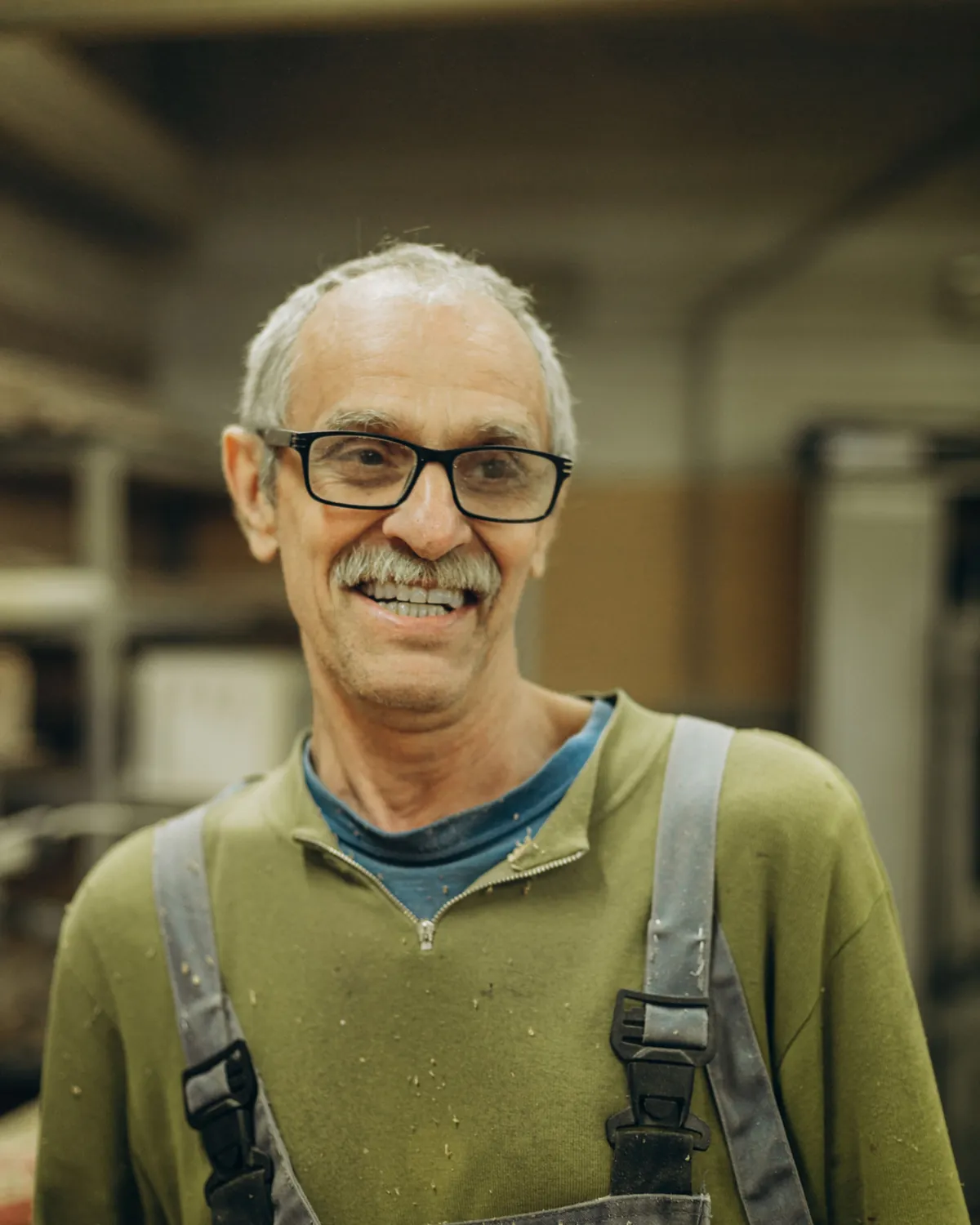 Dennis, smiling, in glasses and work overalls in the workshop