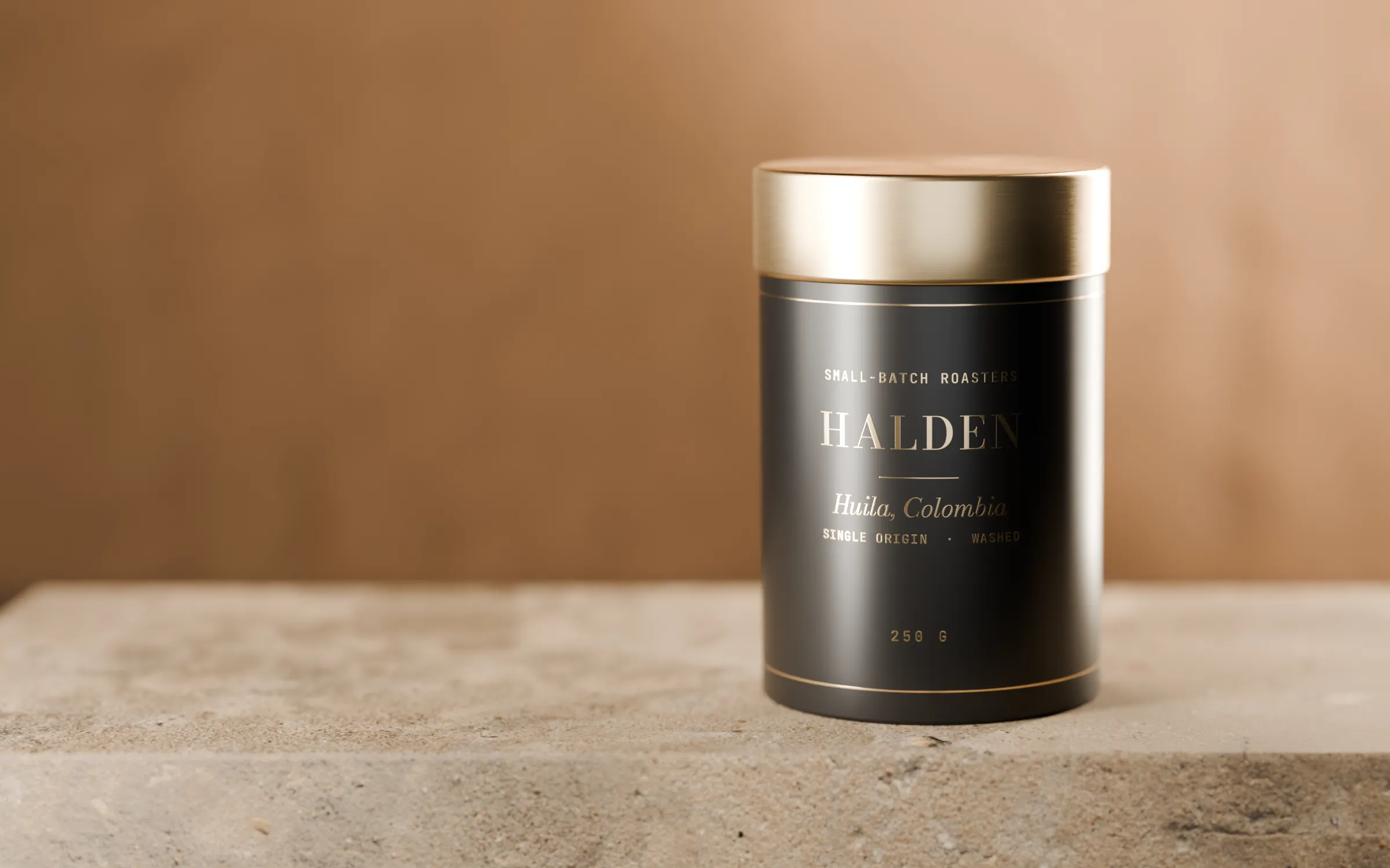 A matte black Halden tin of Huila, Colombia, with a brushed-brass lid, standing on a stone plinth against a warm plaster wall