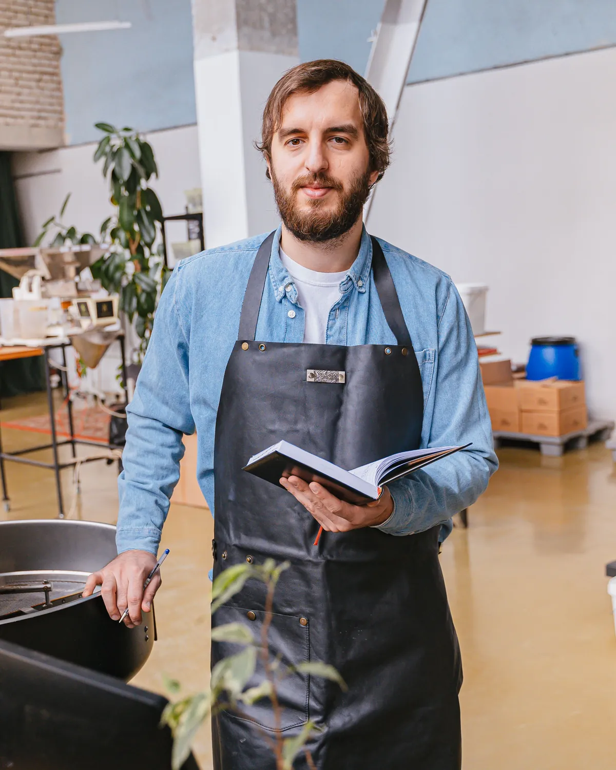 Kit, bearded, in a denim shirt and a dark apron, holding a notebook beside the roaster