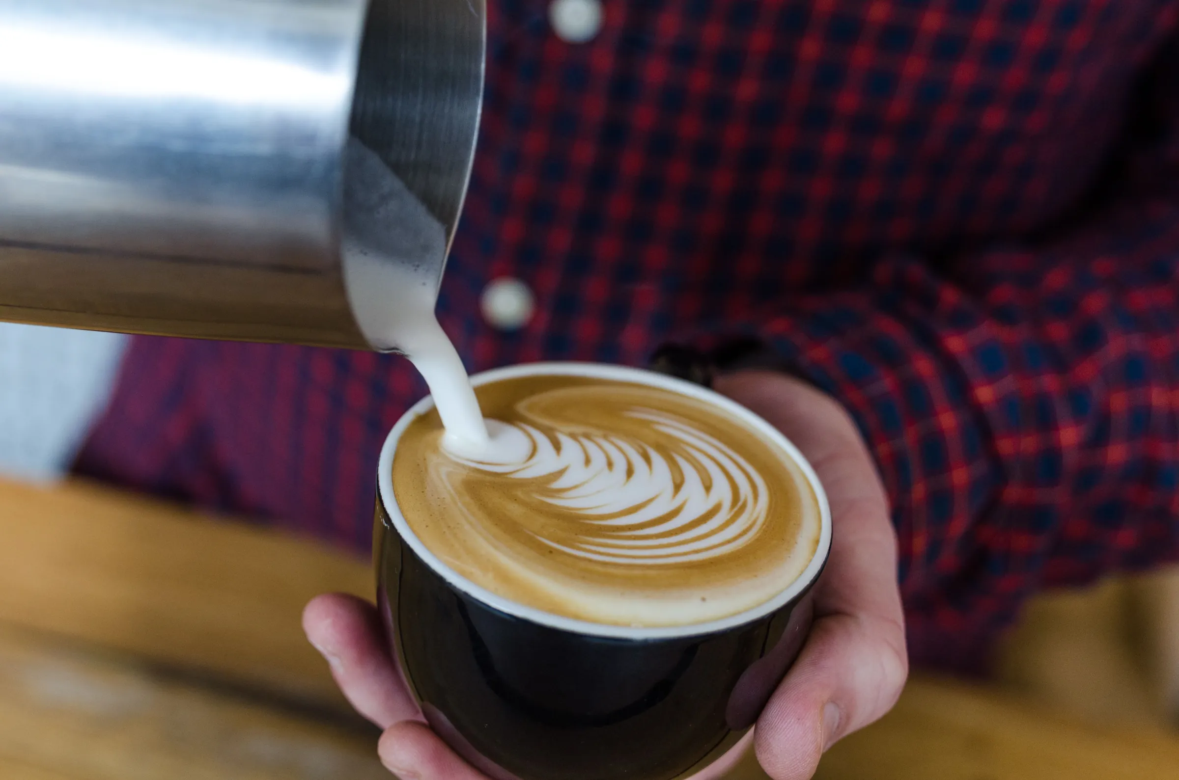 A barista pouring steamed milk into a black cup, a leaf pattern forming in the crema
