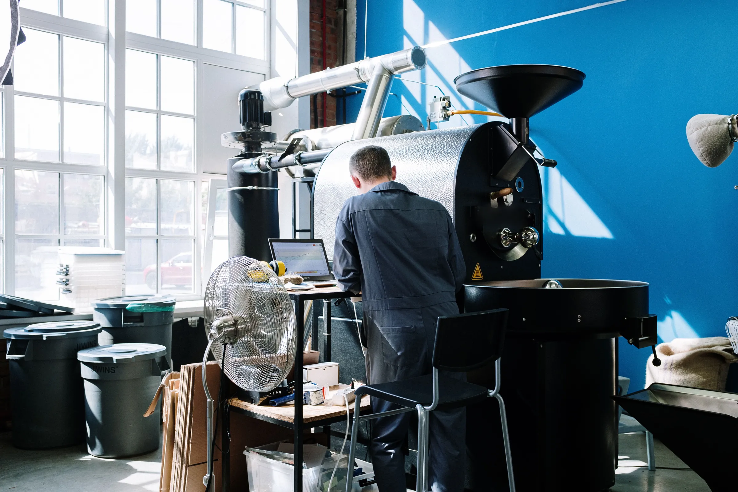 A roaster in overalls working at a black drum roaster in a bright unit with tall windows