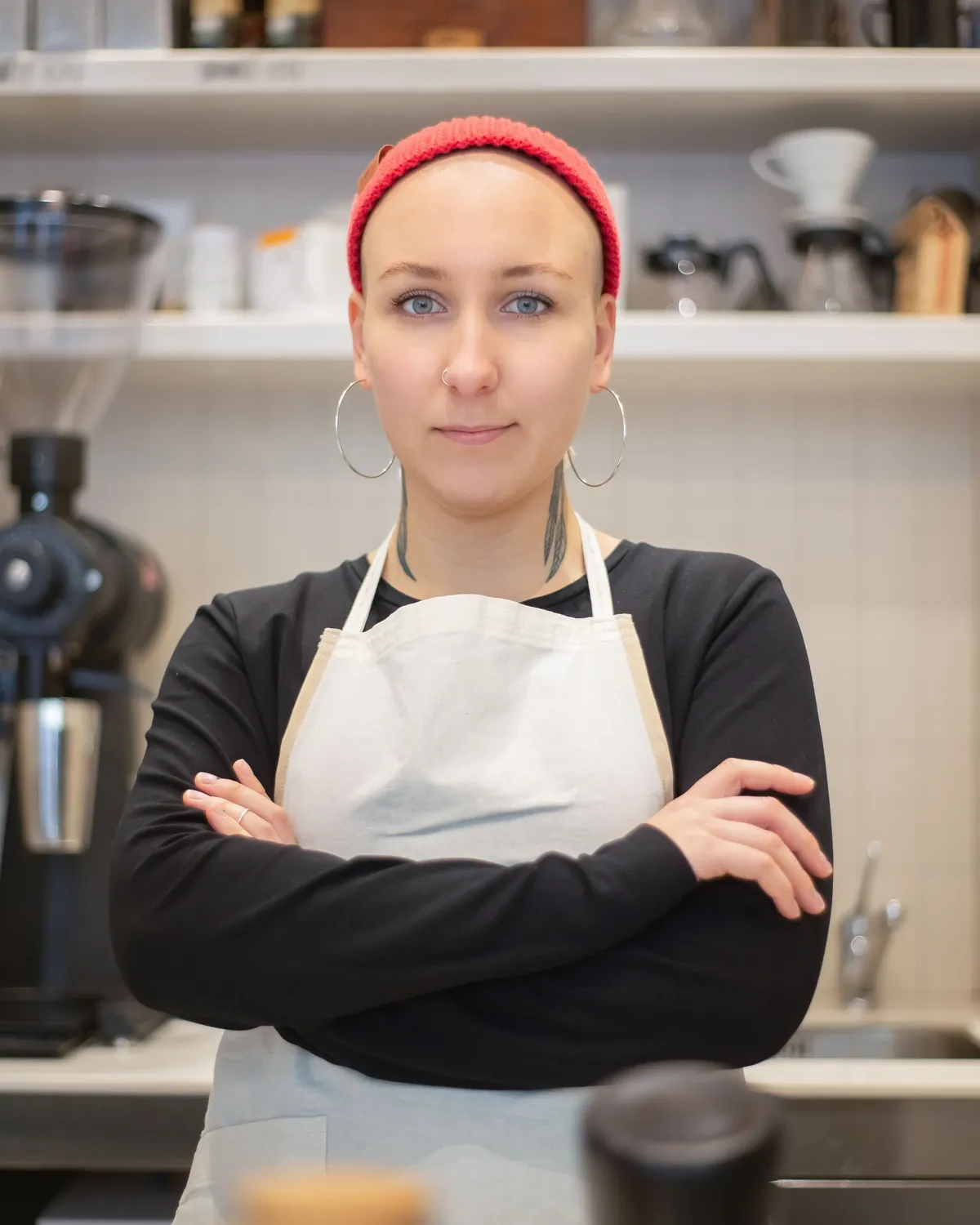 Ruth, in a white apron and a red headband, standing behind a café counter with her arms folded
