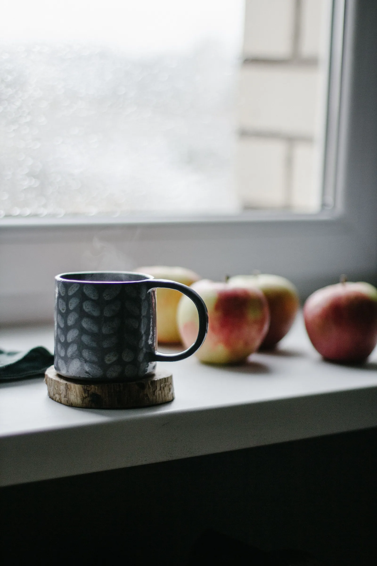 A dark mug of coffee steaming on a white windowsill beside a row of apples, rain on the glass