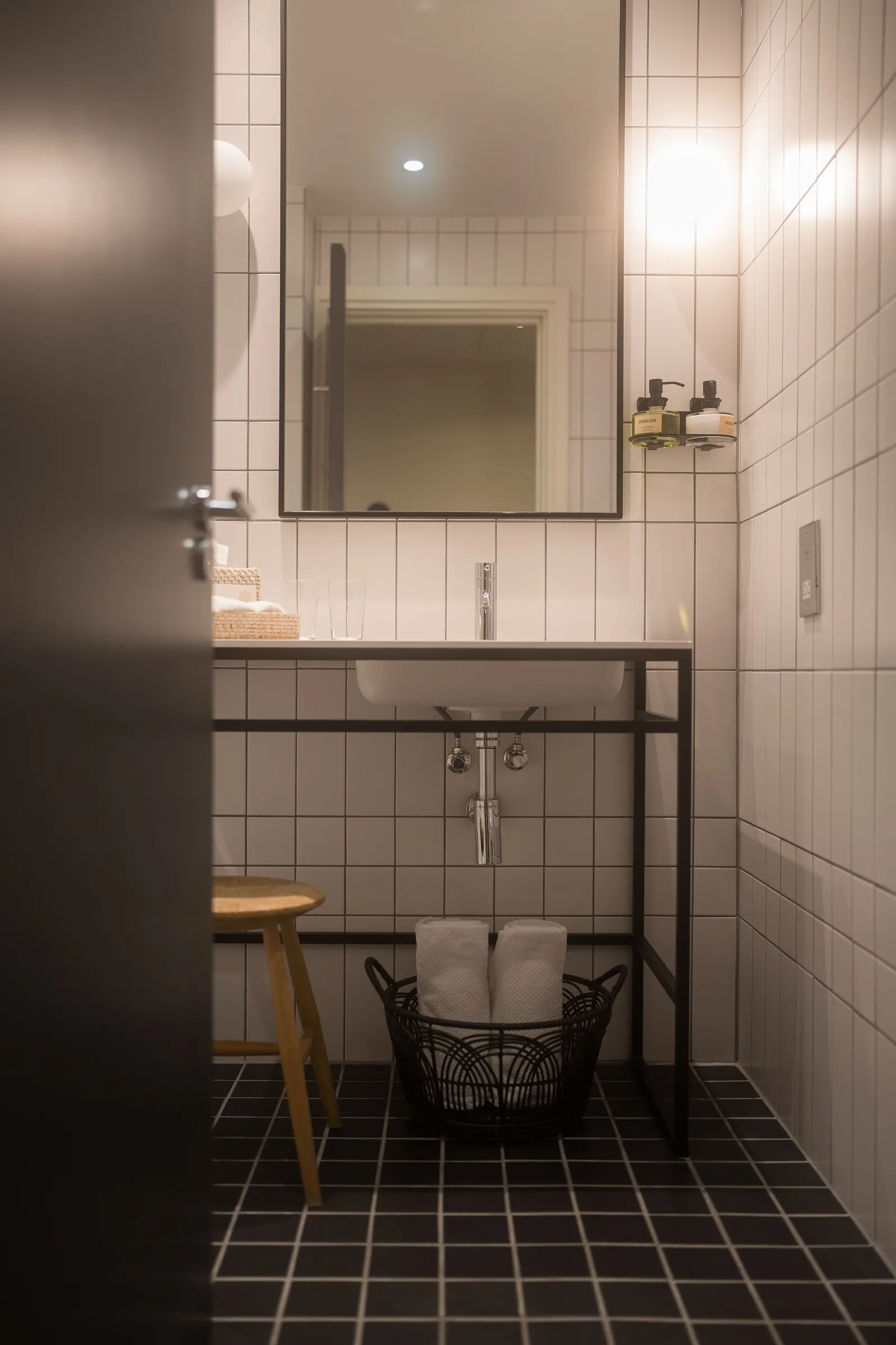 A small bathroom with dark tiles, a wooden stool and a round mirror