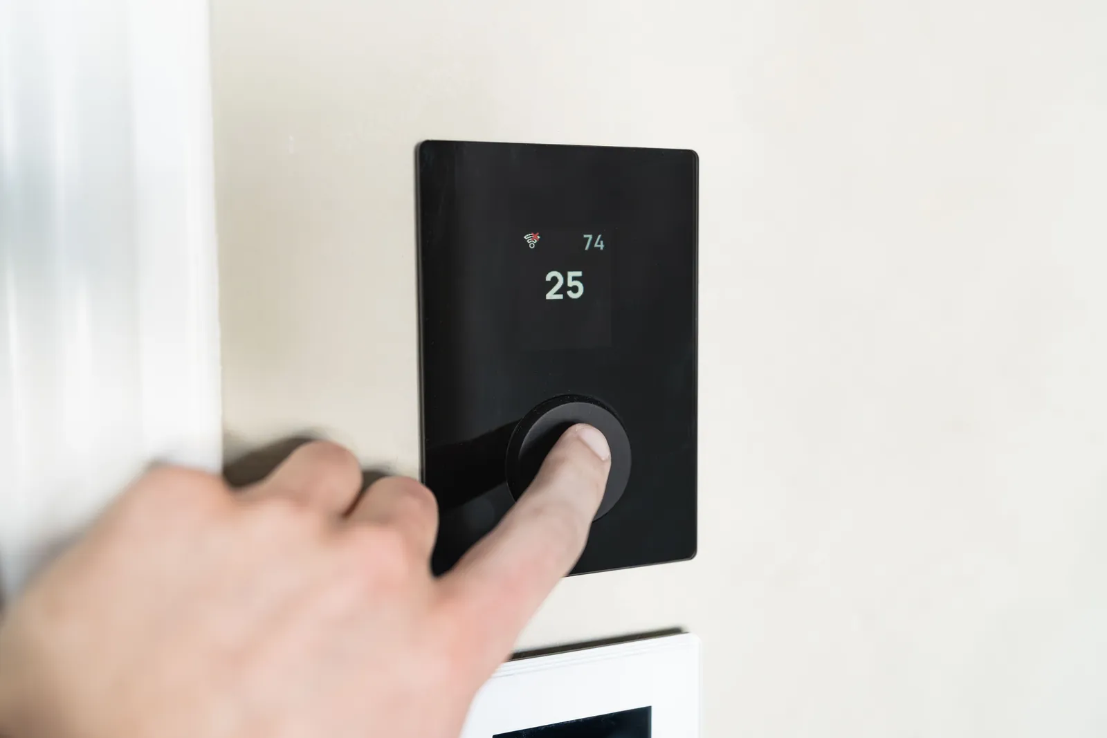 A hand pressing the dial on a black smart thermostat mounted on a white wall