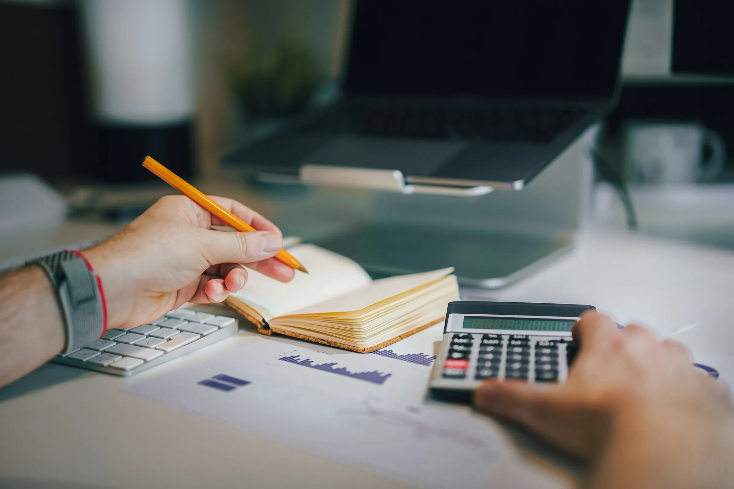 A hand working a desk calculator beside an open notebook and a printed chart