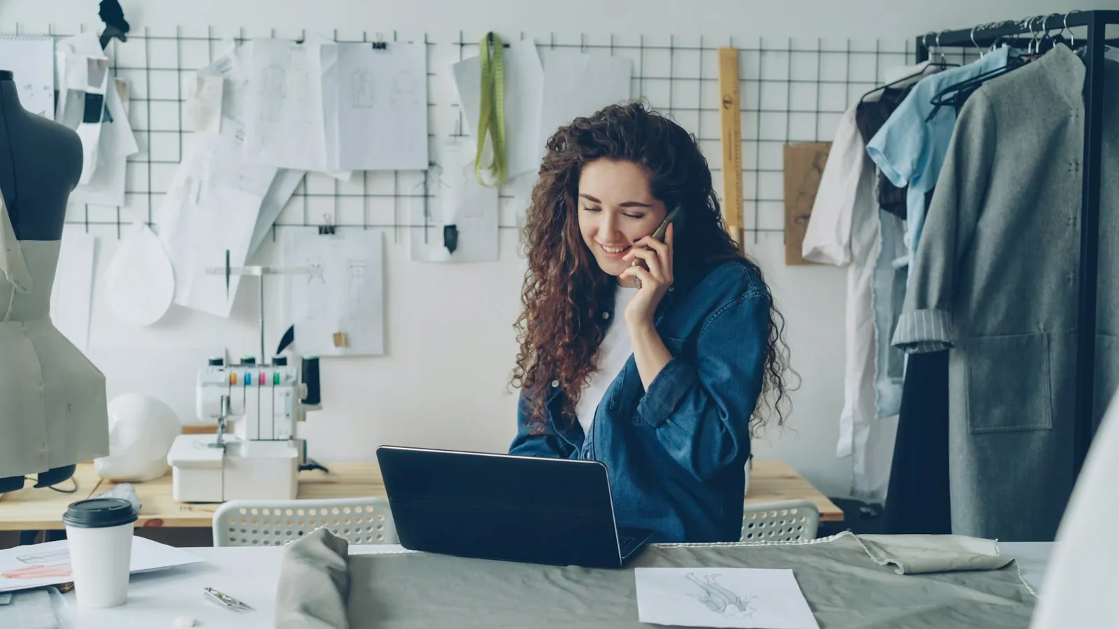 A designer on the phone at her workbench, laptop open, sketches pinned behind her