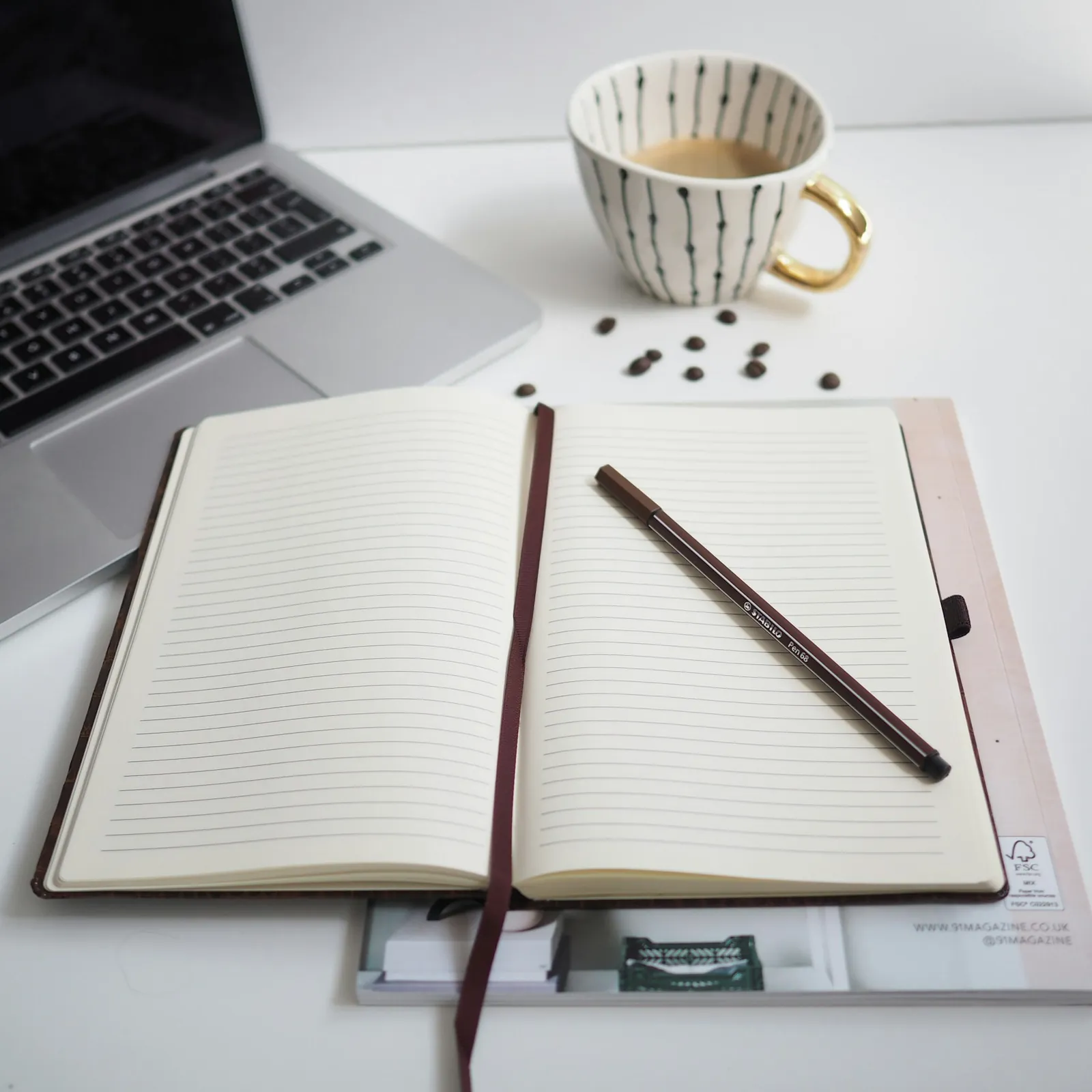 An open notebook and pen beside a laptop, with a coffee