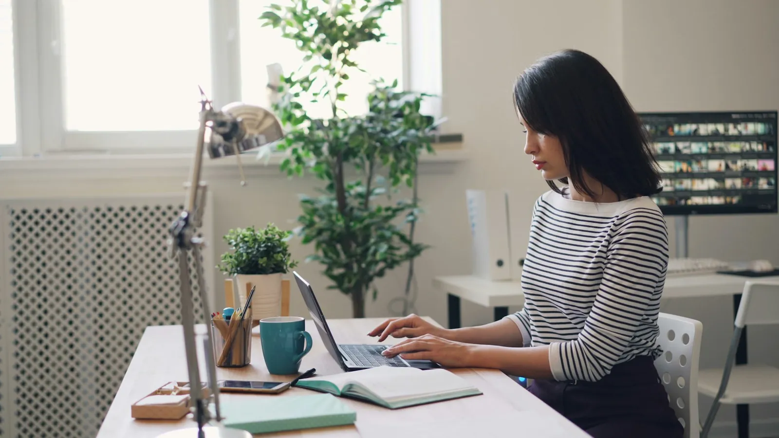 A woman working at a laptop beside a tall window, plants on the desk