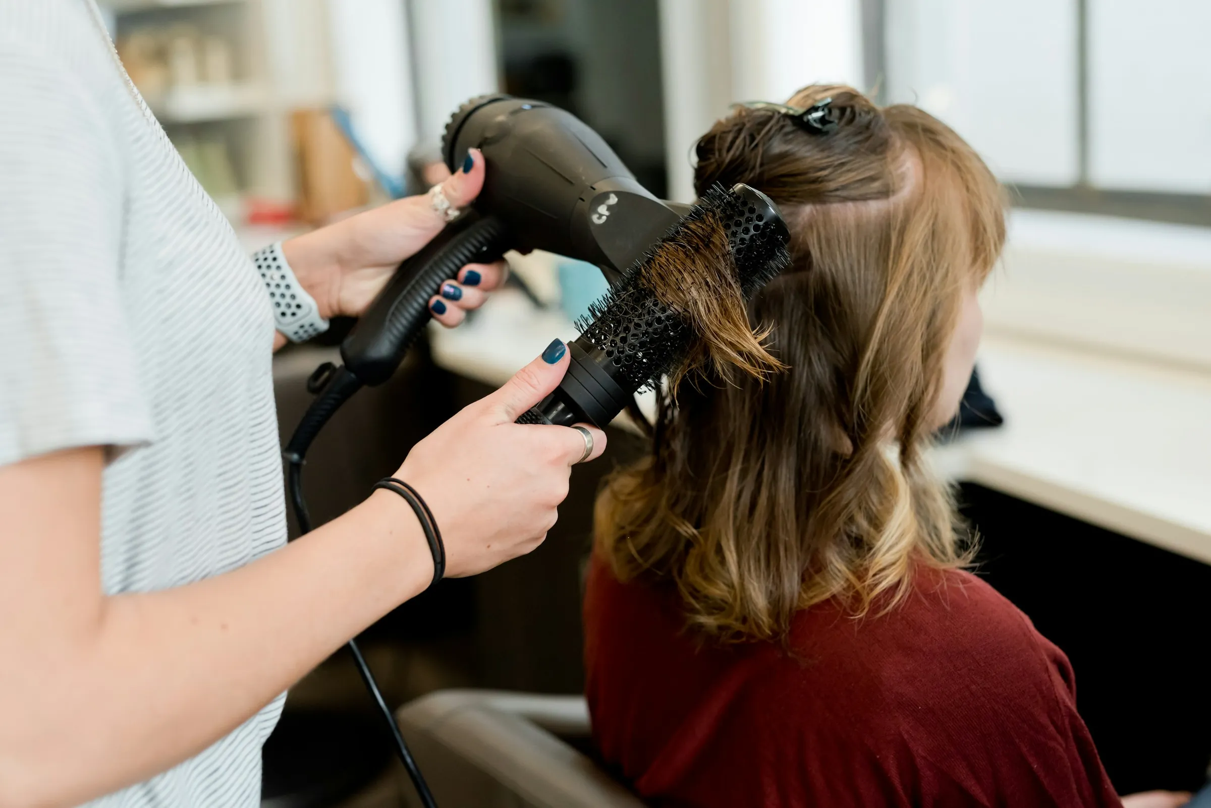 A stylist blow-drying a client's hair with a round brush at the window