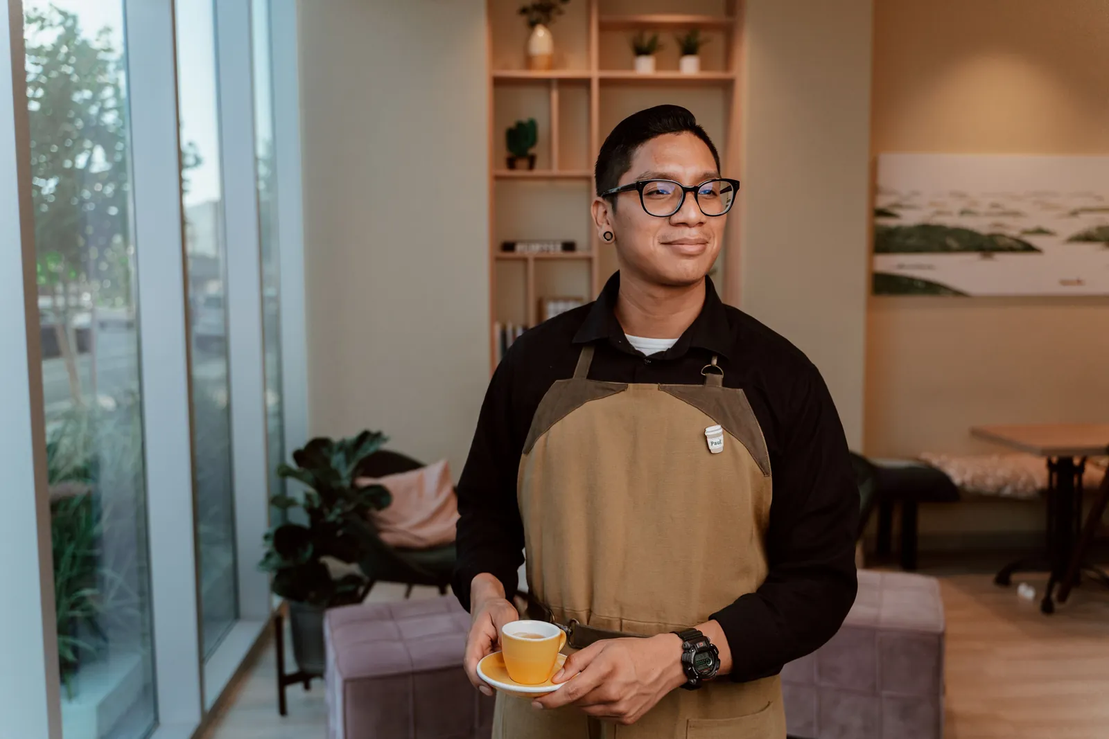 Dan in a tan apron holding a yellow cup, standing by the window
