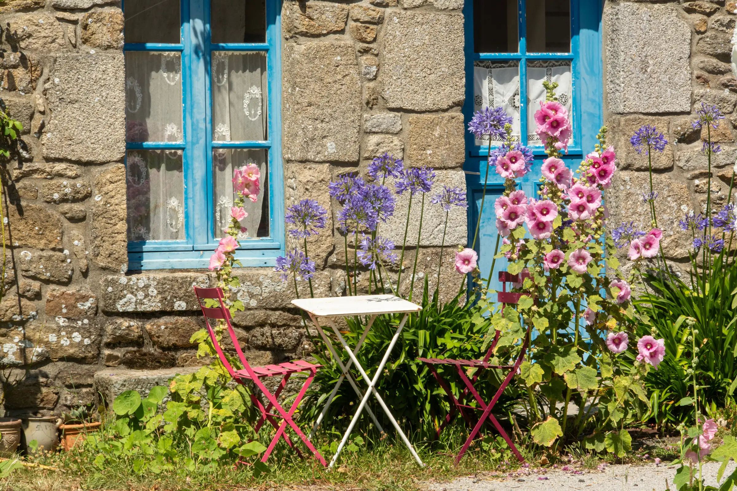 A bistro table and chairs against a stone cottage wall, hollyhocks in flower