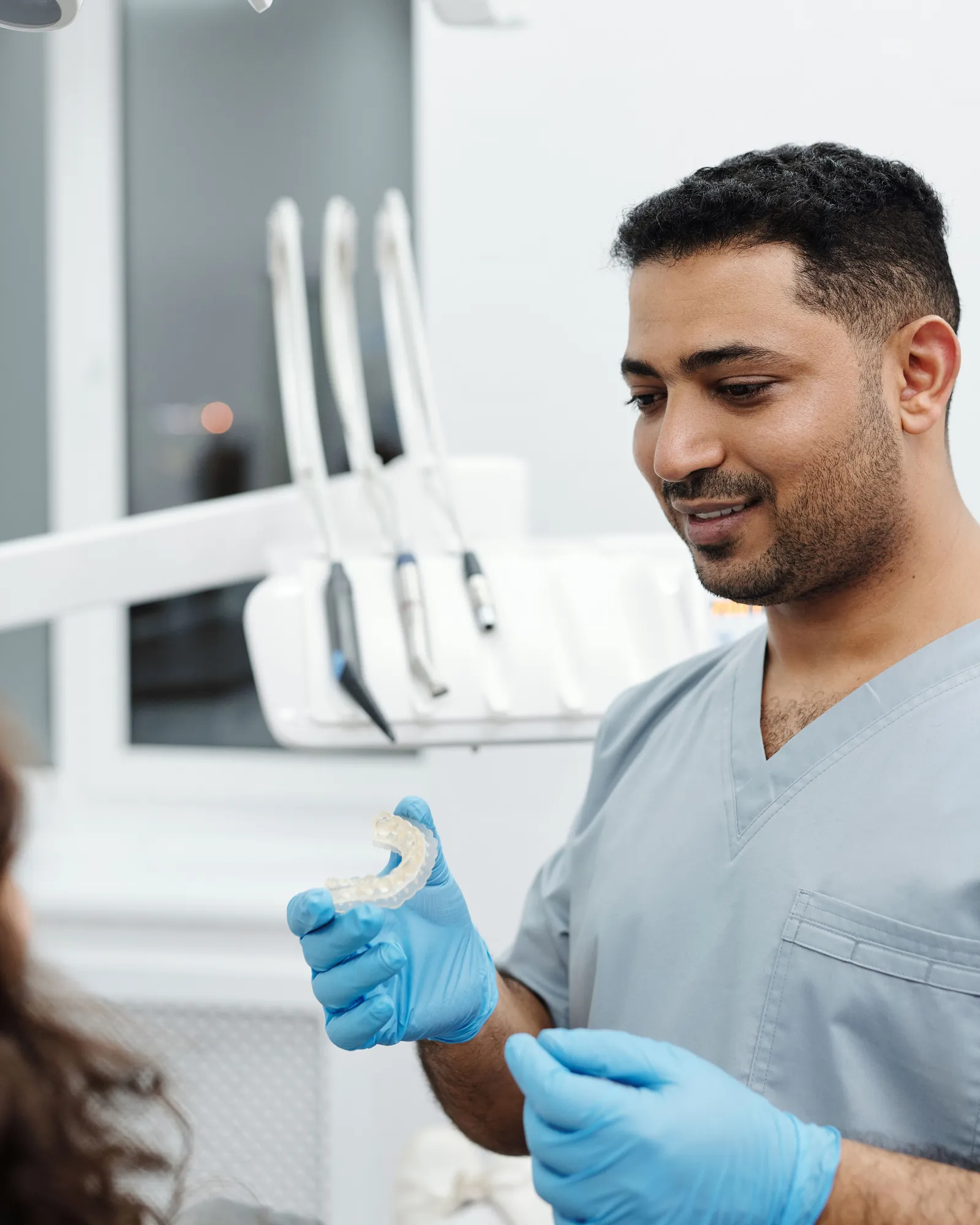 A dentist in grey scrubs holding a clear aligner up in blue-gloved hands