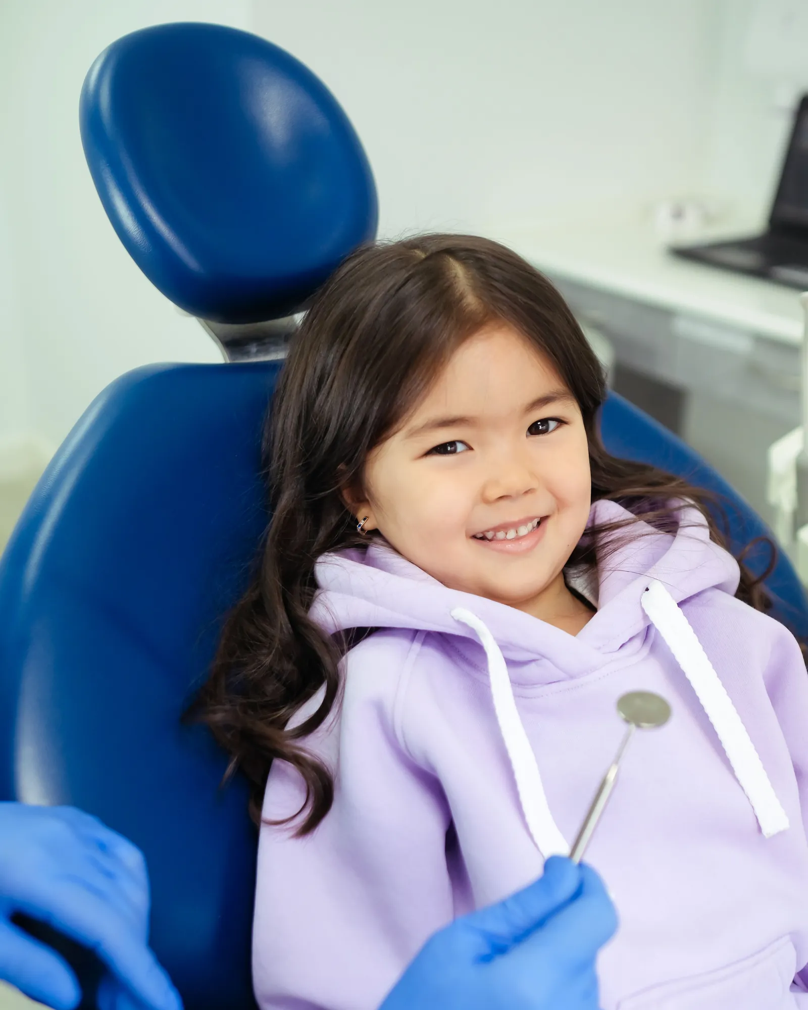 A small girl in a lilac hoodie grinning in a blue dental chair