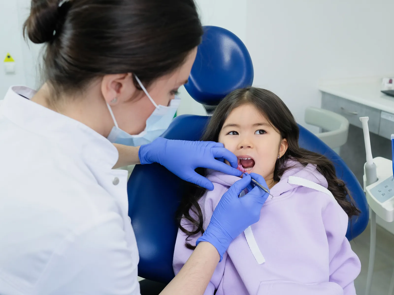 A dentist in white checking a small girl&rsquo;s teeth with a mirror while she sits in a blue chair