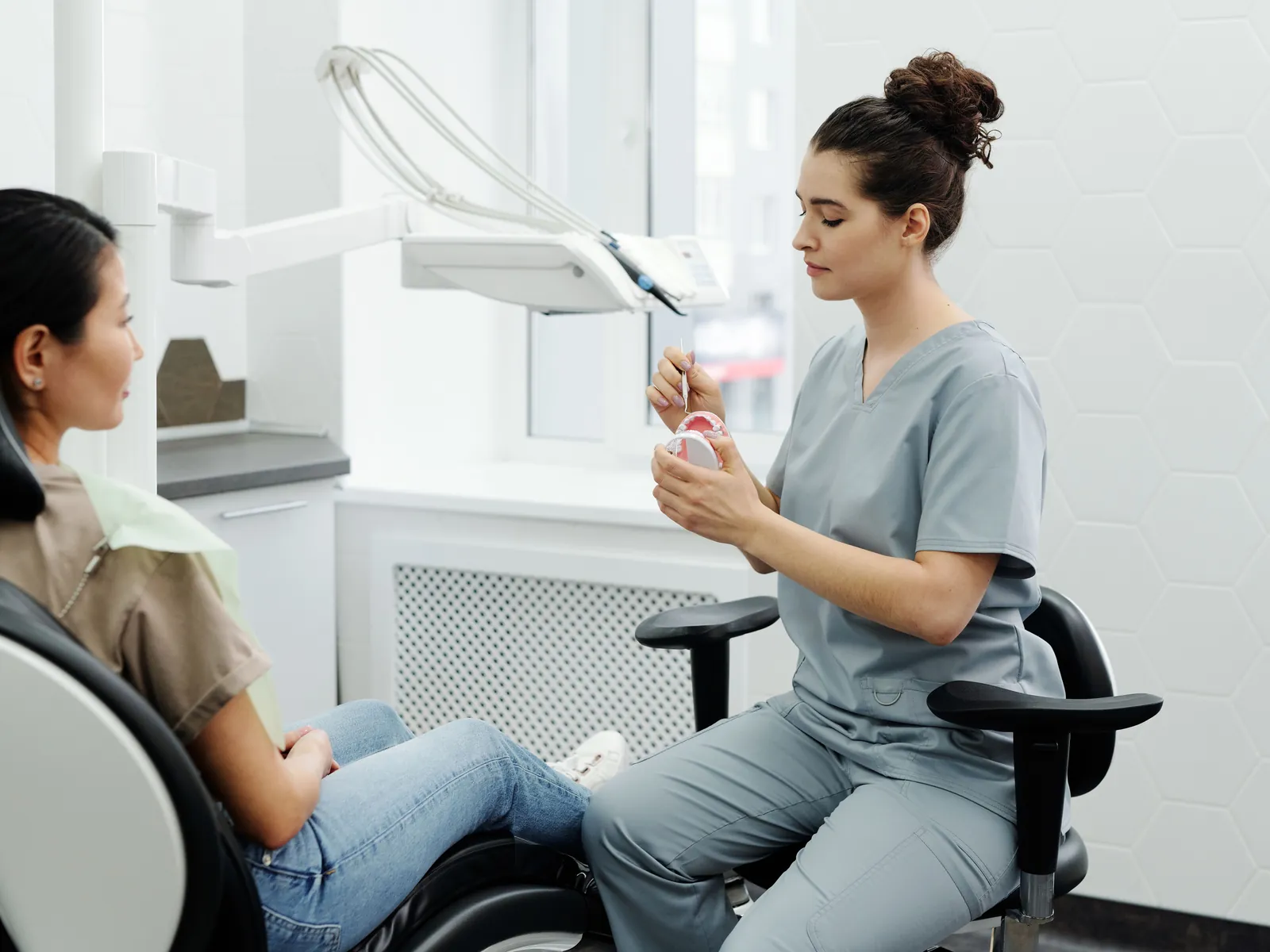 A dentist in grey scrubs sitting beside a patient in the chair, talking rather than treating