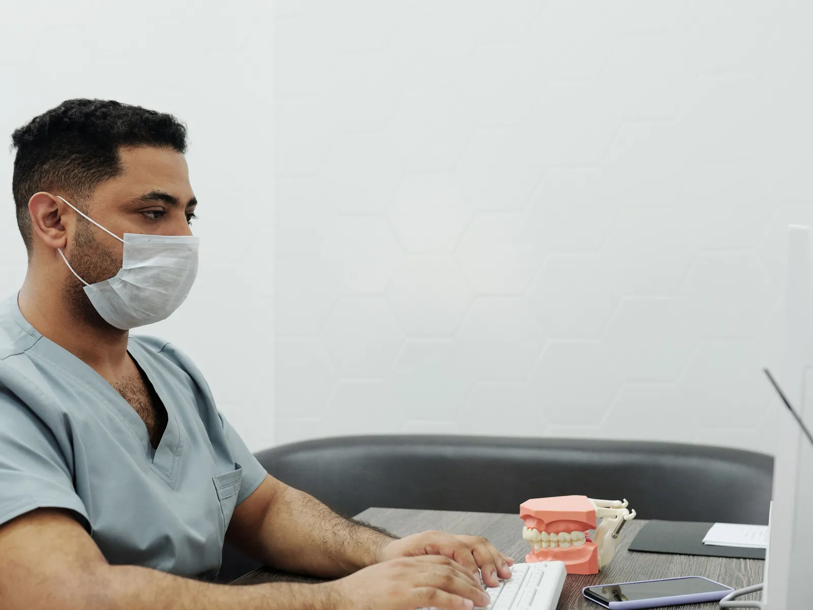 A masked dentist in grey scrubs typing notes at a desk with a set of model teeth beside the keyboard