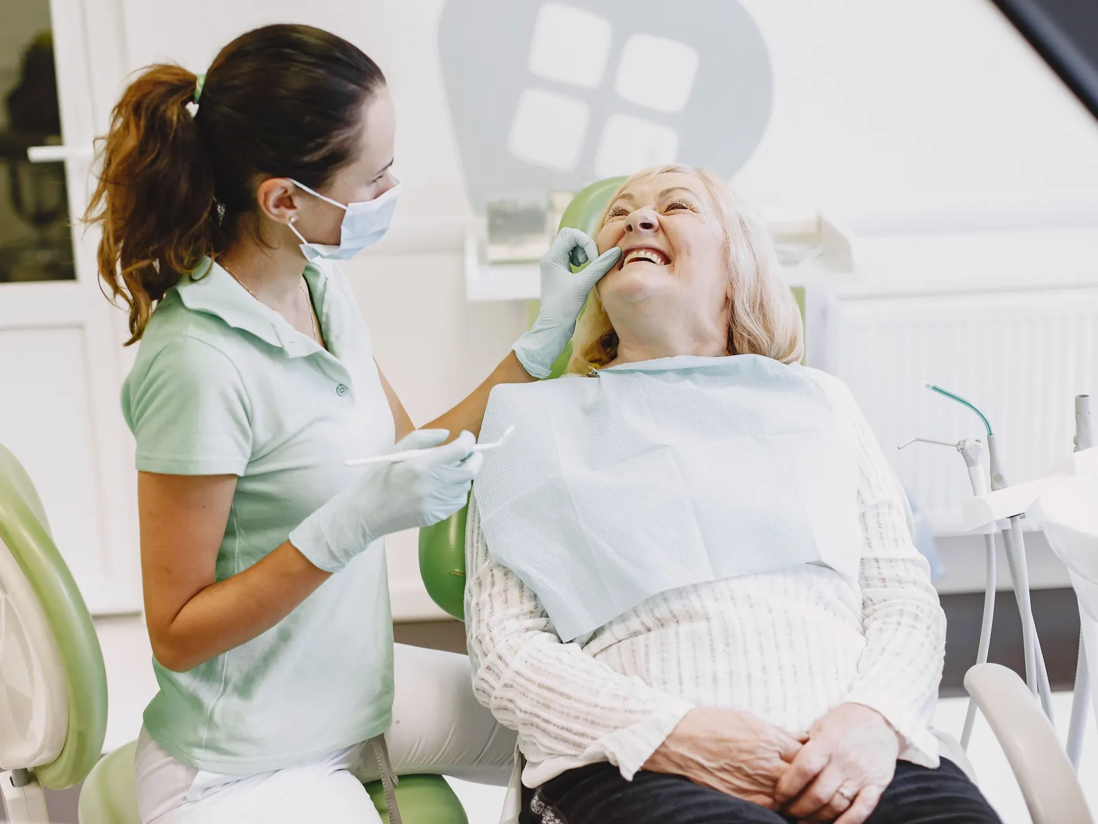 A hygienist in pale green scrubs cleaning the teeth of an older woman who is laughing in the chair