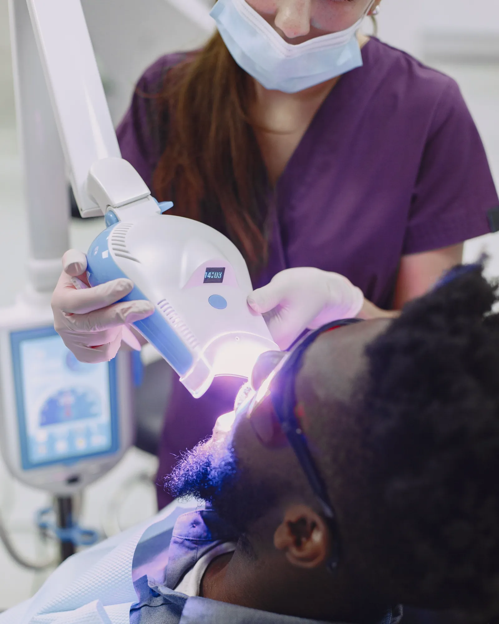 A dentist in purple scrubs holding a blue curing light over a patient's teeth
