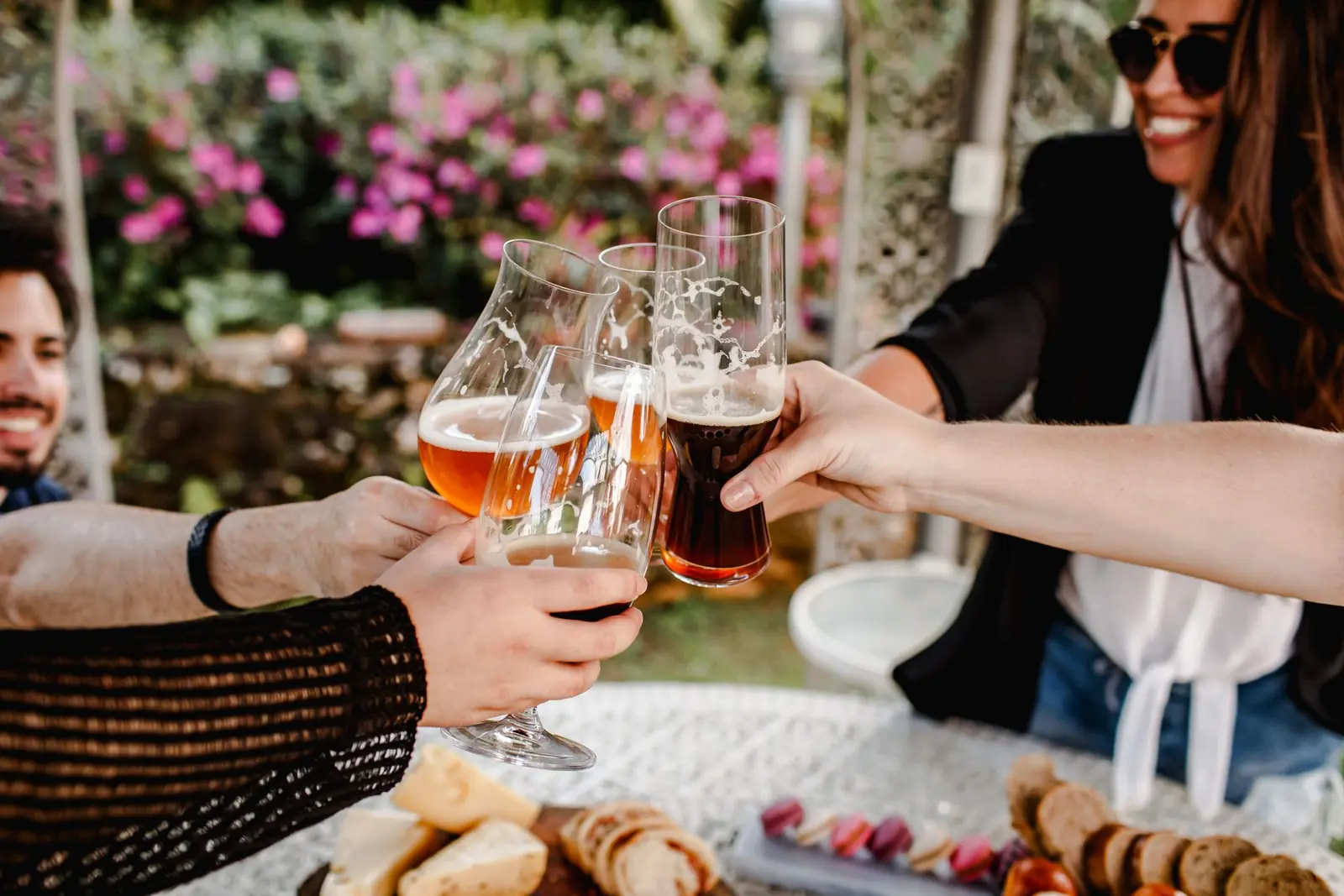 Two people touching glasses together over a table outdoors, with flowers behind them.