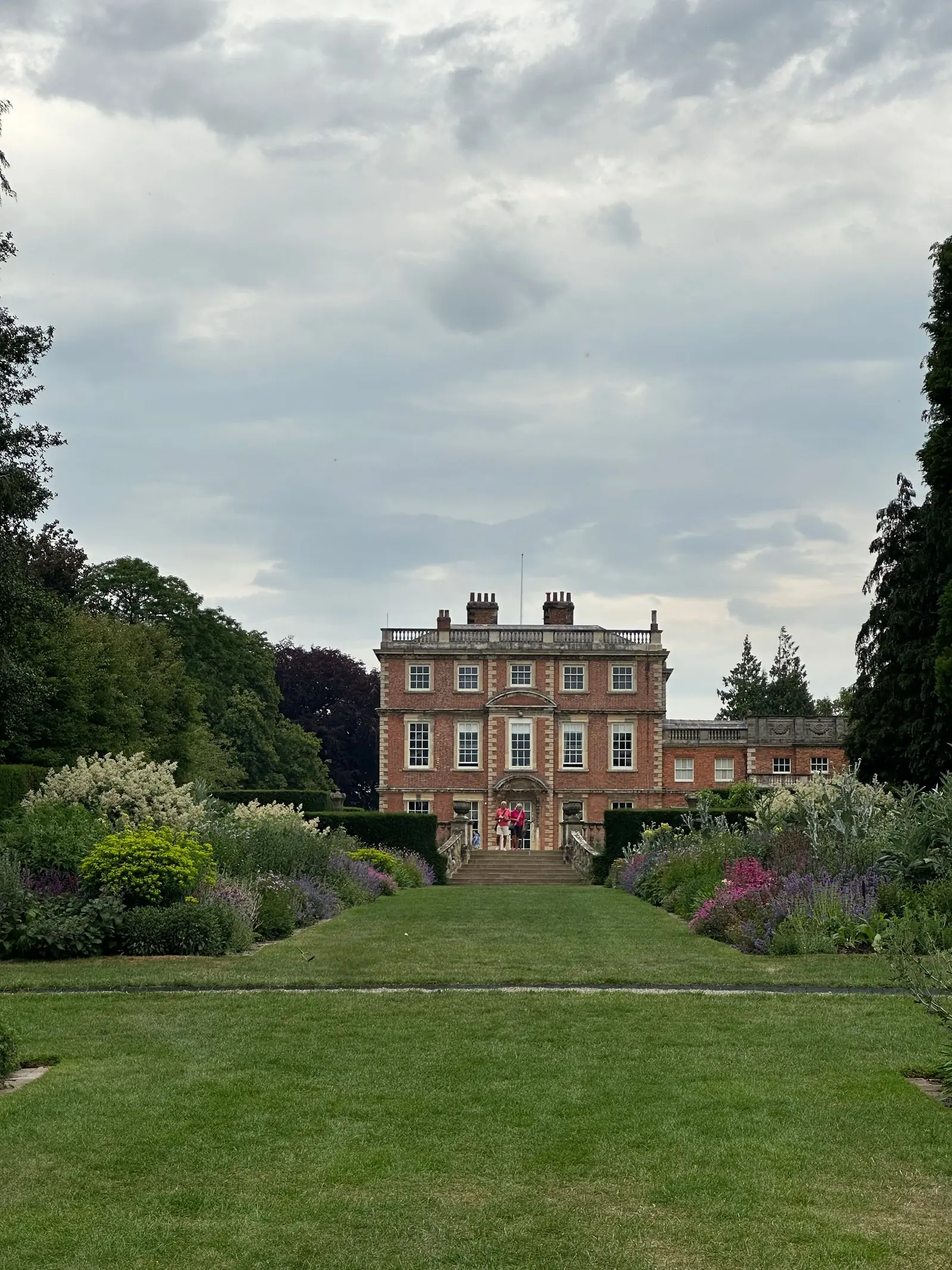 A red-brick Georgian house behind deep planted borders and a striped lawn, under a grey English sky.