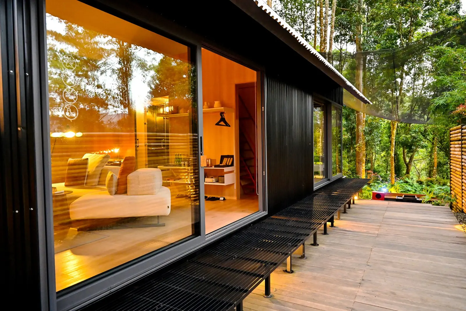 A dark timber-clad garden building at dusk, its full-height glass doors open and warm light spilling onto a timber deck.