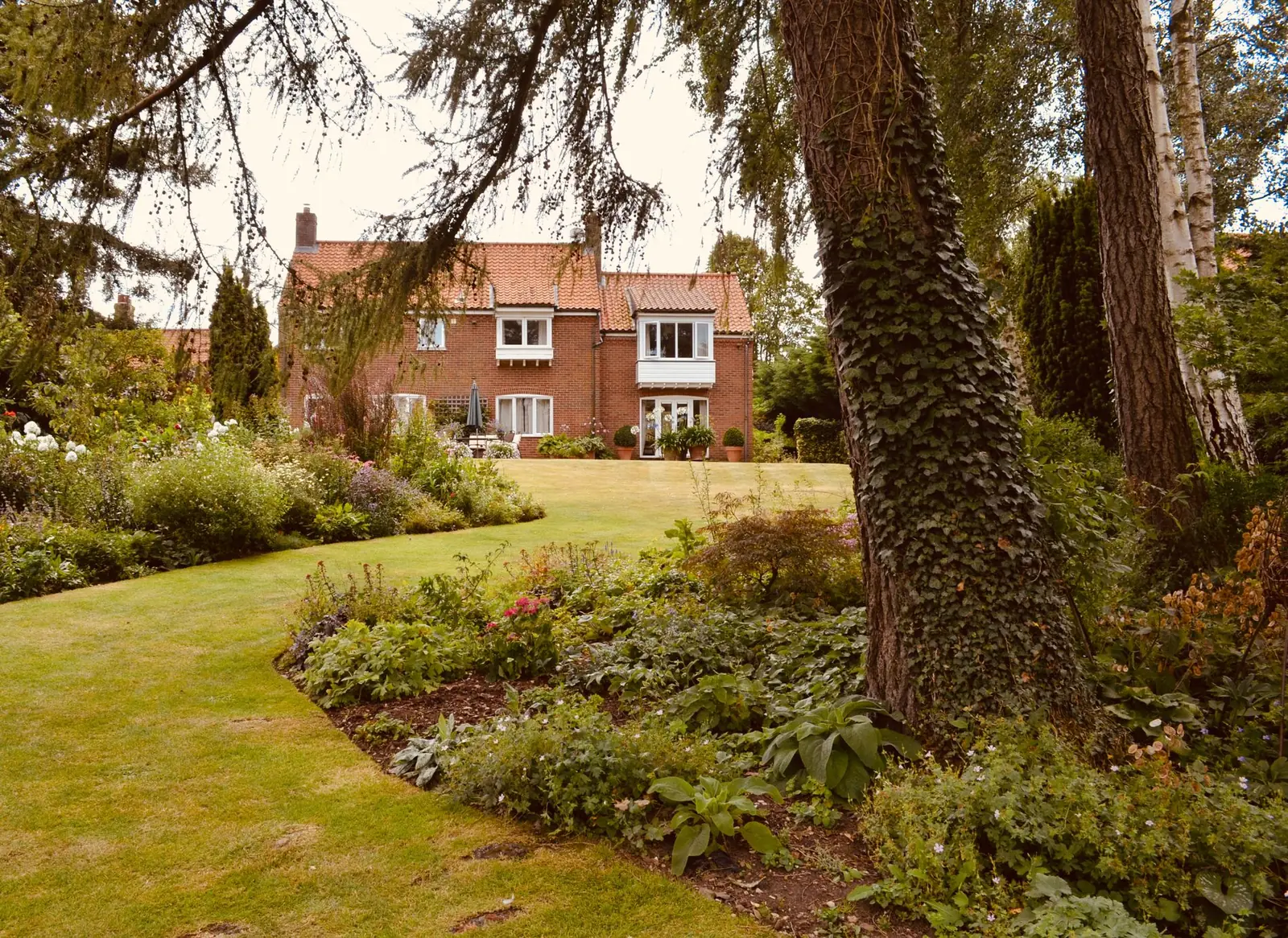 A red-brick house behind a lawn and deep planted borders, with a mature conifer in the foreground.