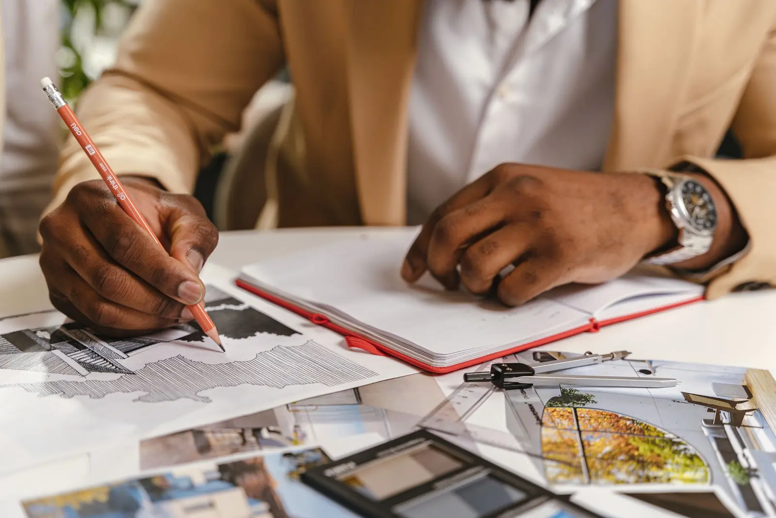 Hands drawing with a pencil on a plan, with material samples and photographs spread across the table.