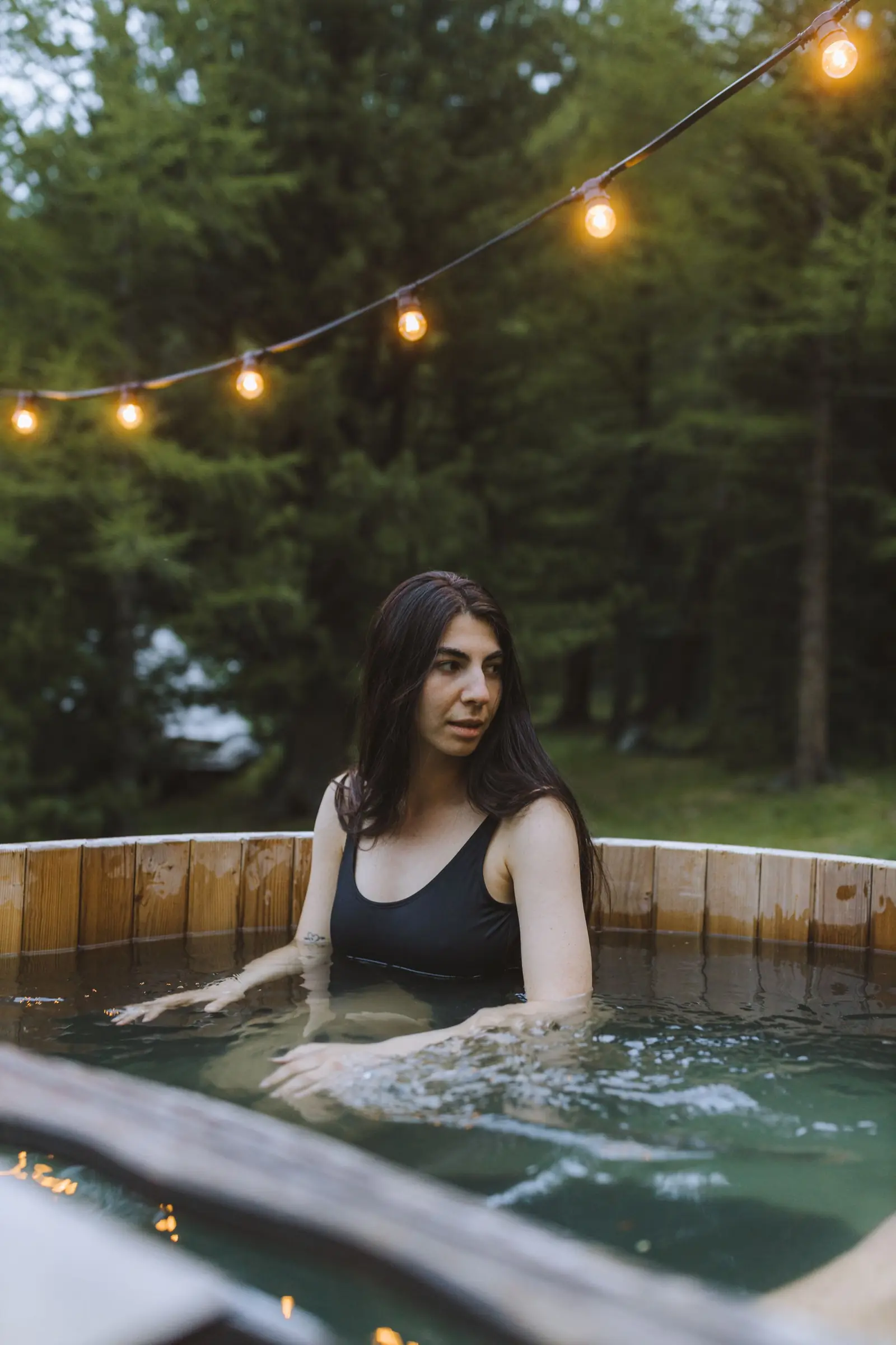 A woman in a black swimming costume sitting in a round wooden hot tub at dusk, a string of warm bulbs hung above her and dark trees behind.