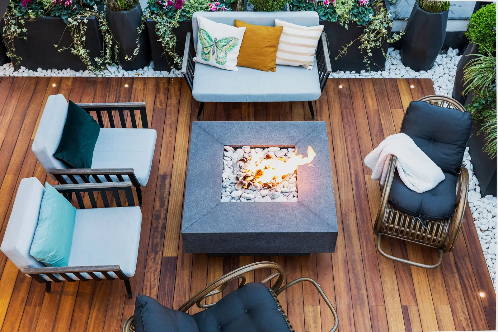 A timber deck at night with low seating, a lit fire table and planting along the boundary.