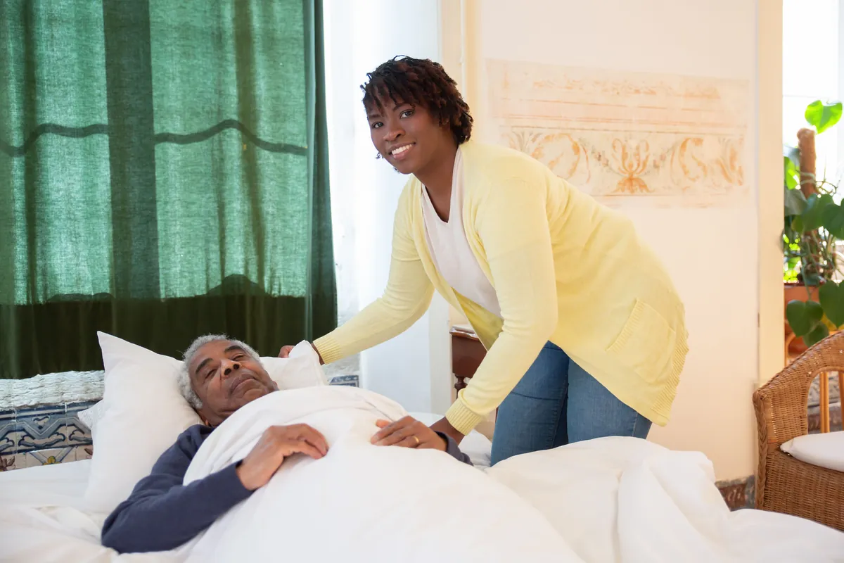 A carer in a yellow cardigan tucking in a resident's bed in a bright room.