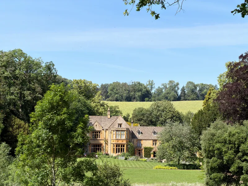 A stone house with gables set back behind a wide lawn and mature trees.