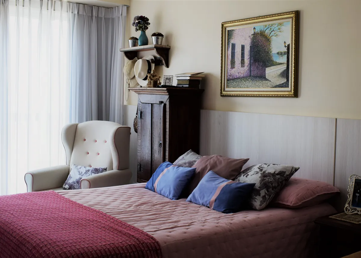 A bedroom with a wing-backed armchair, a made bed with cushions and a painting above the headboard.