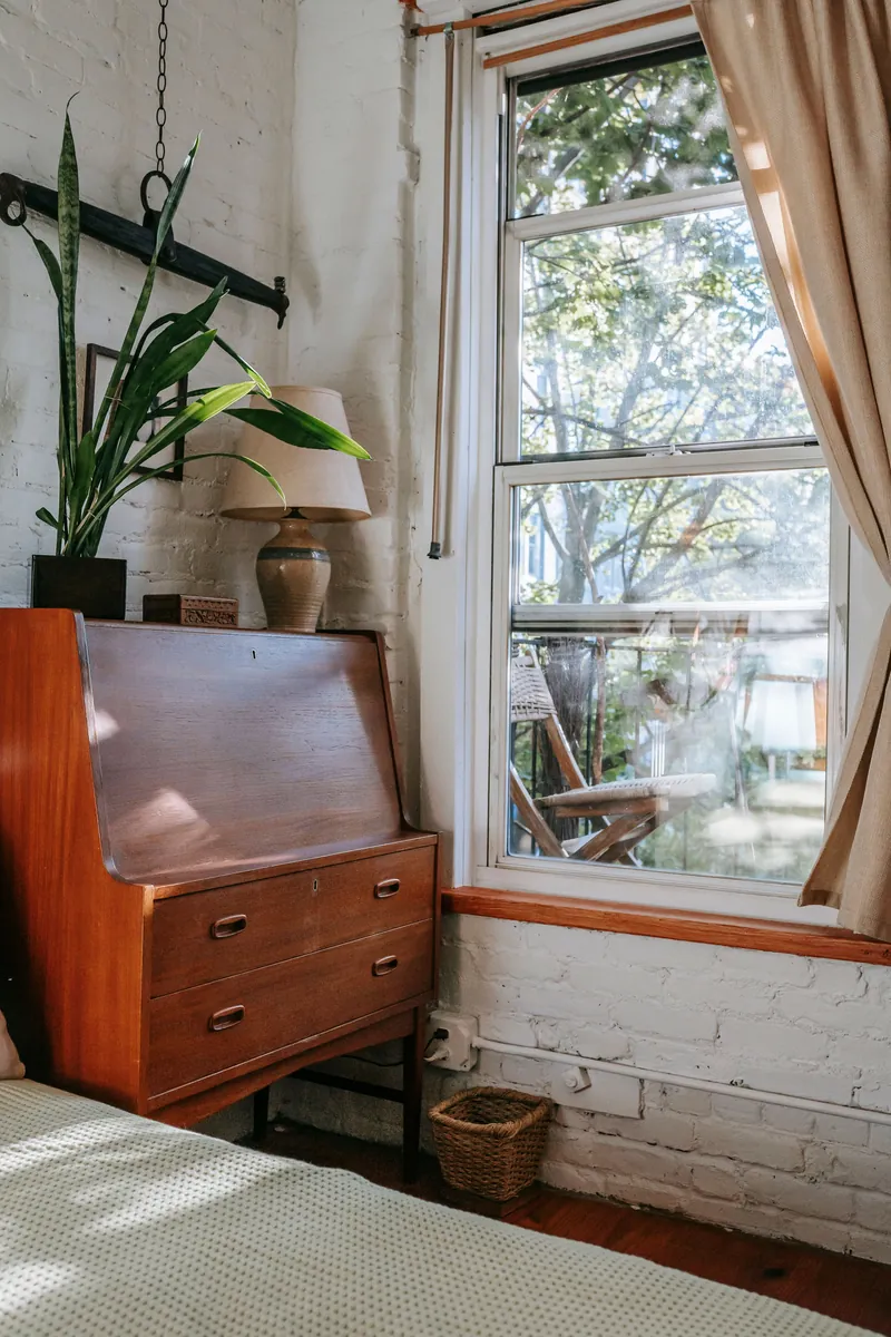 A bedroom corner with a wooden bureau, a lamp and a plant beside a bright window.