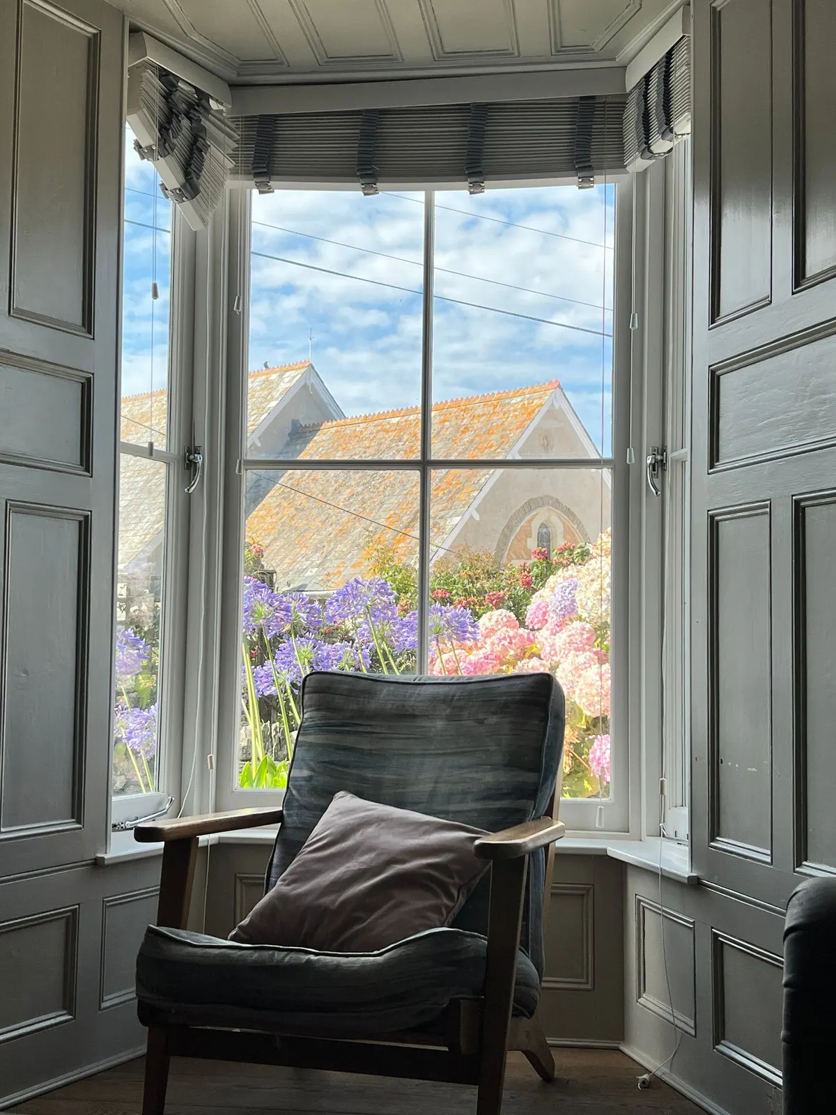 A chair at an open window looking out over hydrangeas and a stone roof.